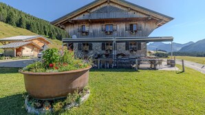 Un bâtiment rustique en bois, doté d’un espace extérieur abrité pour s’asseoir, entouré de verdure et de montagnes.