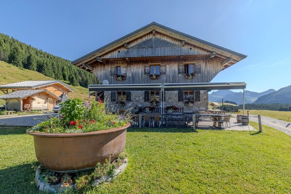 Un bâtiment rustique en bois, doté d’un espace extérieur abrité pour s’asseoir, entouré de verdure et de montagnes.