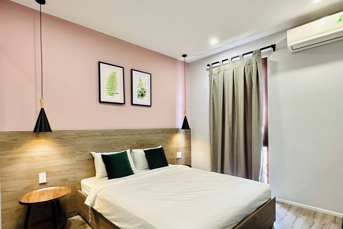 A modern bedroom with a wooden headboard, a bed with white bedding, two bedside lamps, and a small wooden stool.