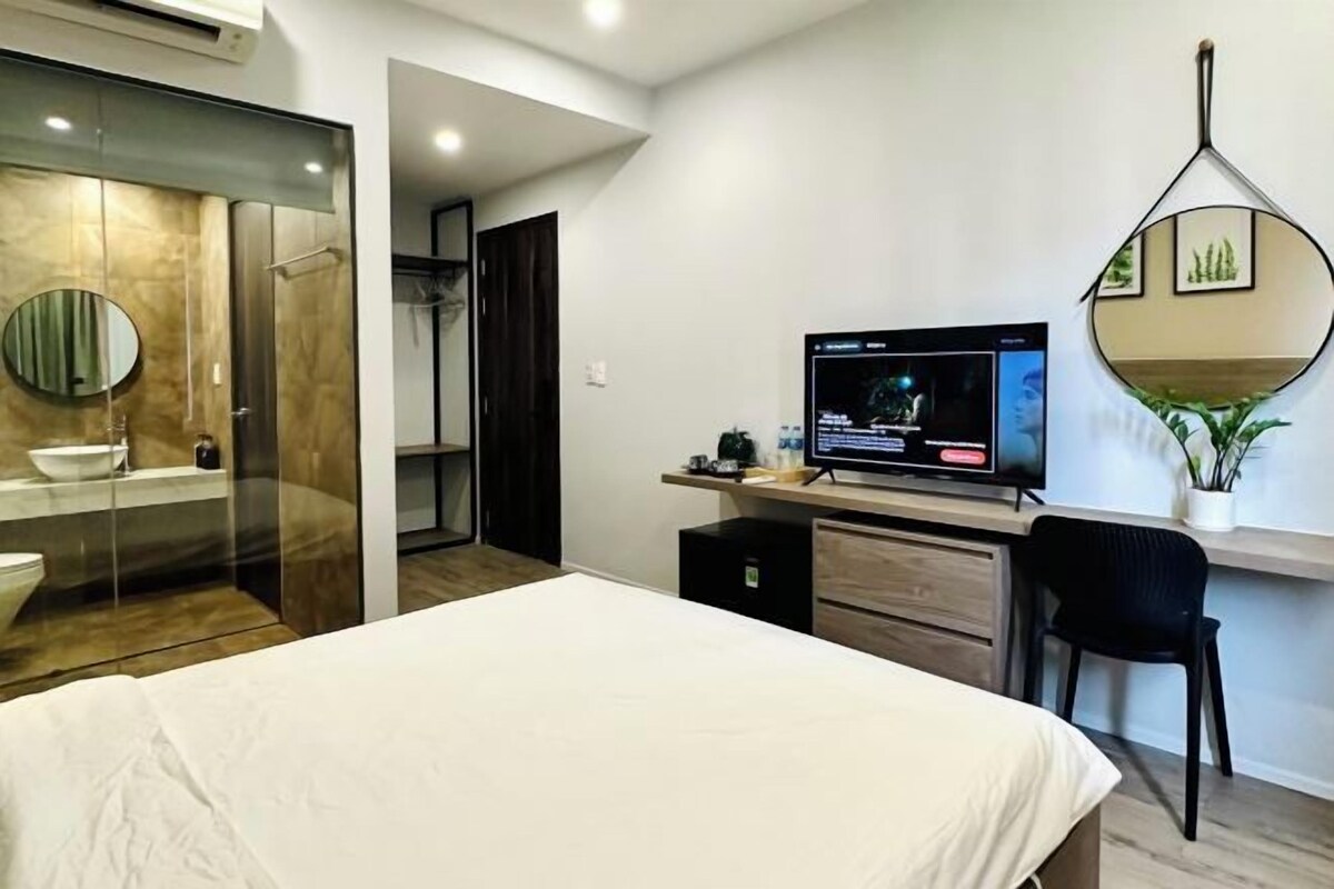 A modern hotel room with a bed, a desk with a computer, a TV, a mirror, and a bathroom with a glass partition.