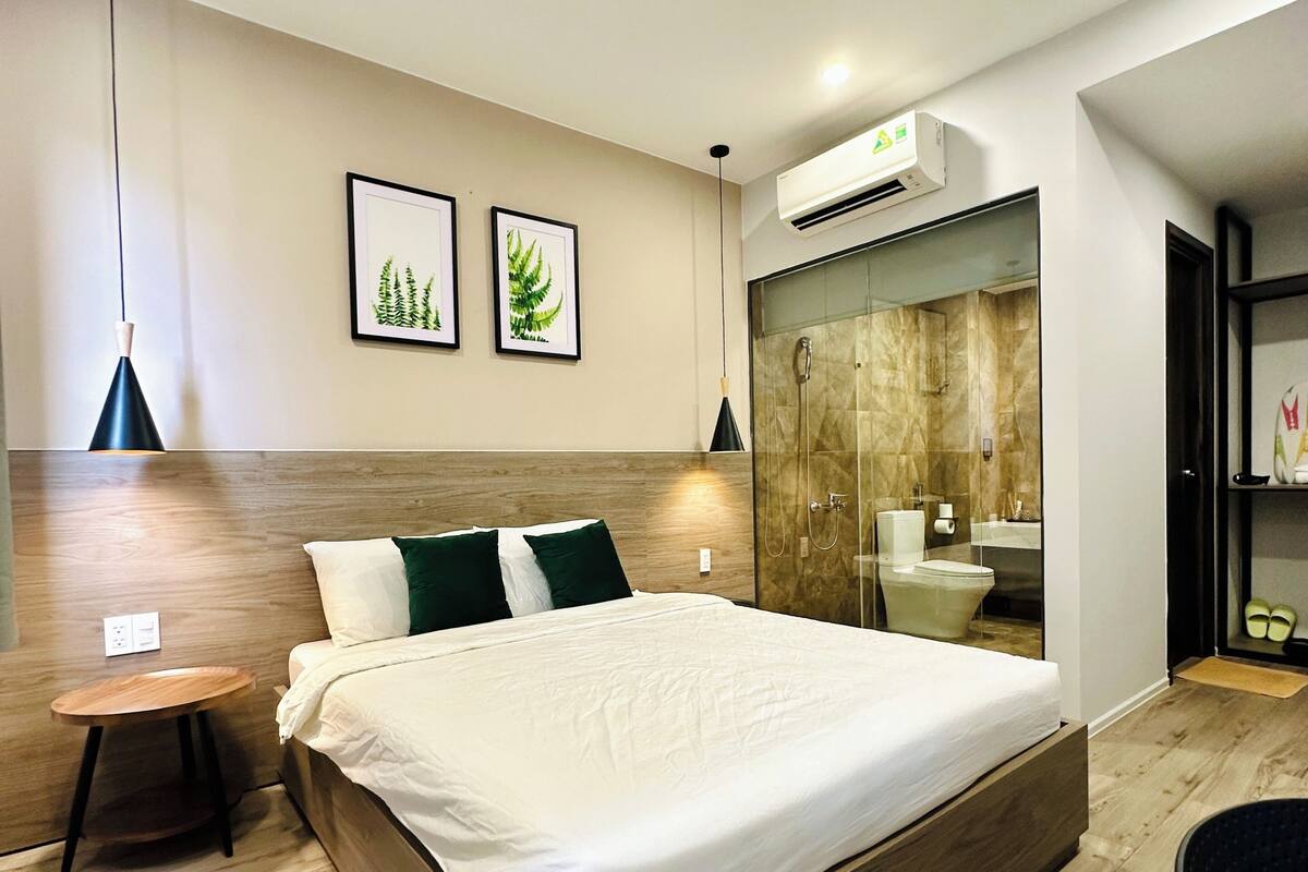 A modern bedroom with a large bed, bedside tables, a hanging light, and a bathroom with a glass shower enclosure.
