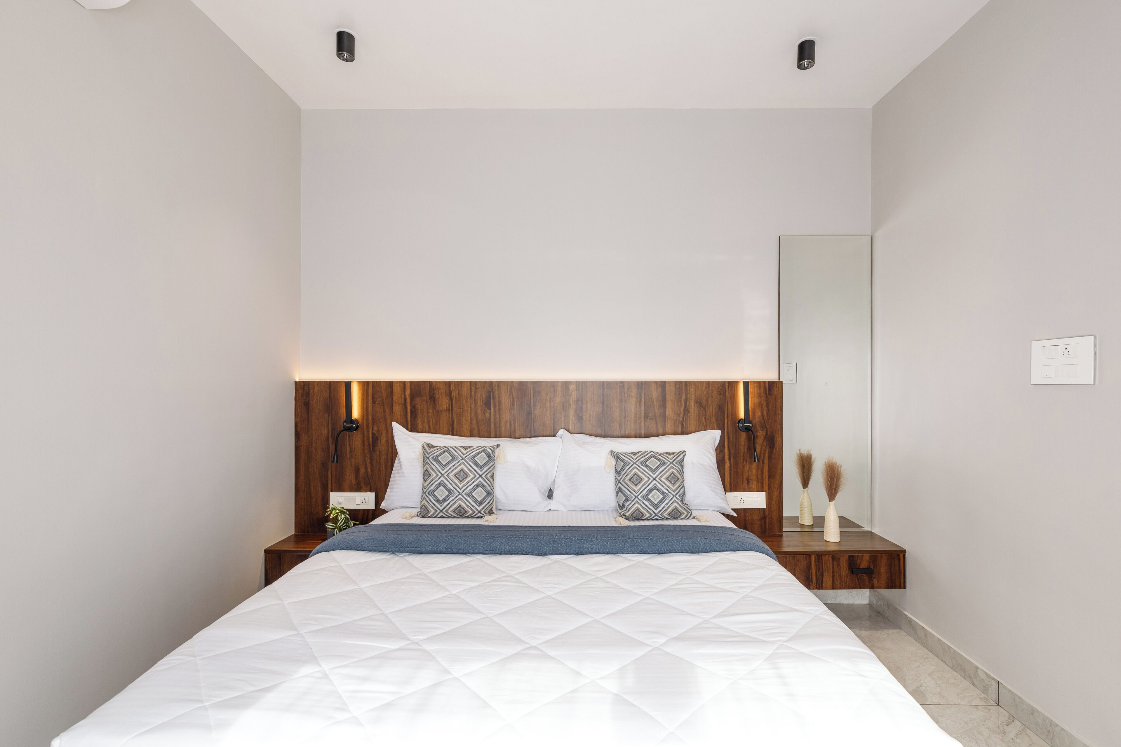 Un dormitorio moderno con cabecera de madera, una cama grande con ropa blanca, dos almohadas estampadas, una mesita de noche con un florero y un buró.