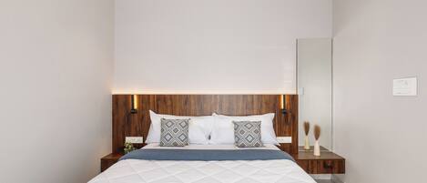 Un dormitorio moderno con cabecera de madera, una cama grande con ropa blanca, dos almohadas estampadas, una mesita de noche con un florero y un buró.