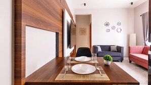 A dining area with a wooden table set for two, a television mounted on the wall, and a living room with a sofa and decorative plates. - The Monarch Stay by Red Olive (Bengaluru)