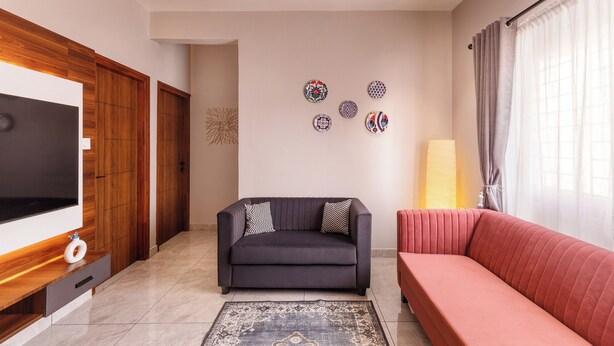 A living room with a dark grey sofa, a red sofa, a flat-screen TV, wooden cabinets, and a patterned rug. - The Monarch Stay by Red Olive (Bengaluru)