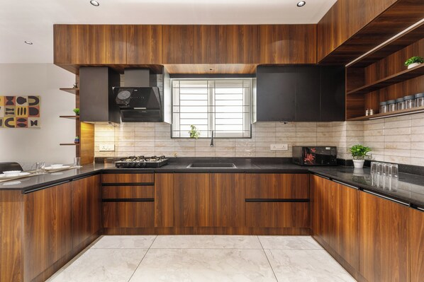 A modern kitchen with wooden cabinets, a black countertop, and a window above the sink. - Aurum Heights by Red Olive (Bengaluru)