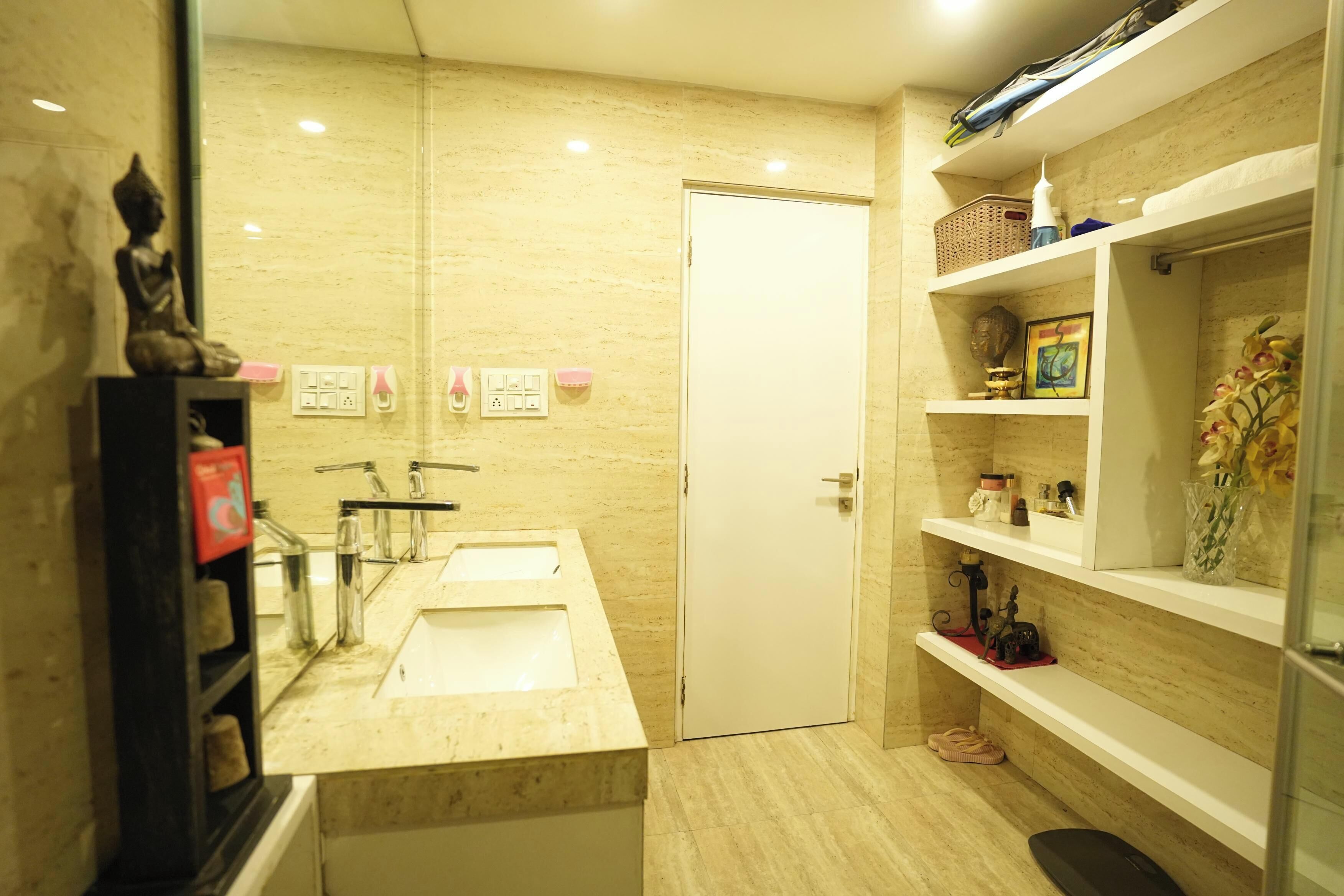 A modern bathroom with two sinks, a toilet, and a shower. There are shelves with toiletries and a statue on the left.