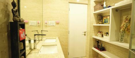 A modern bathroom with two sinks, a toilet, and a shower. There are shelves with toiletries and a statue on the left.