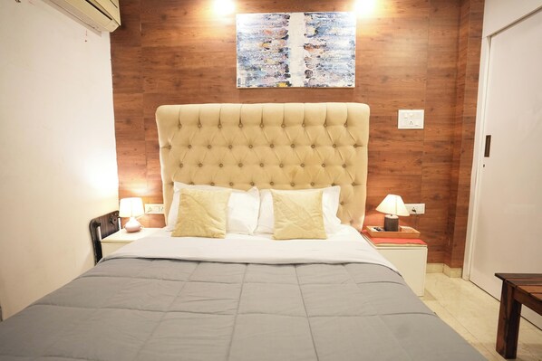 A bedroom with a tufted headboard, a bed with a grey quilt, two bedside tables with lamps, and a wooden wall with a colorful artwork above the bed.