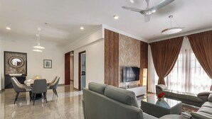 A modern living room with a sofa, a coffee table, and a TV. There is a dining area with a table and chairs. - Emerald Apartments by Red Olive (Bengaluru)