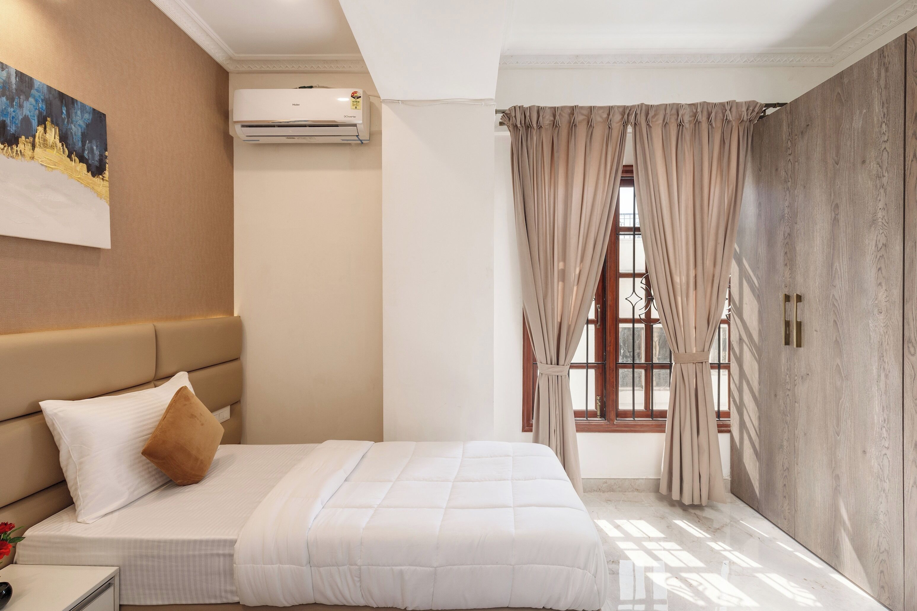 A hotel room with a bed, a nightstand, an air conditioning unit, a window with curtains, and a painting on the wall.