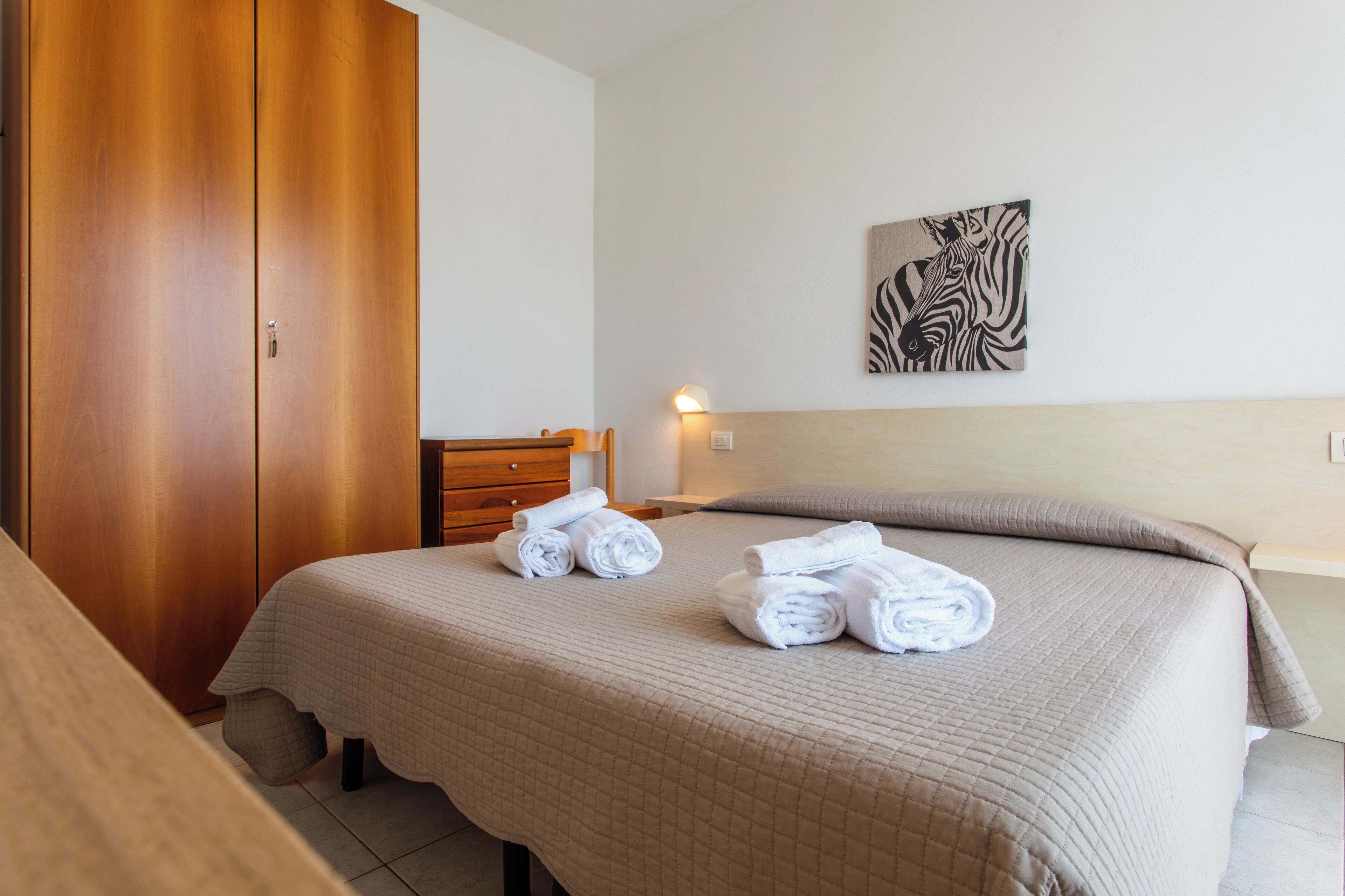 A neatly made bed with rolled towels, a wooden wardrobe, and a zebra artwork on the wall.