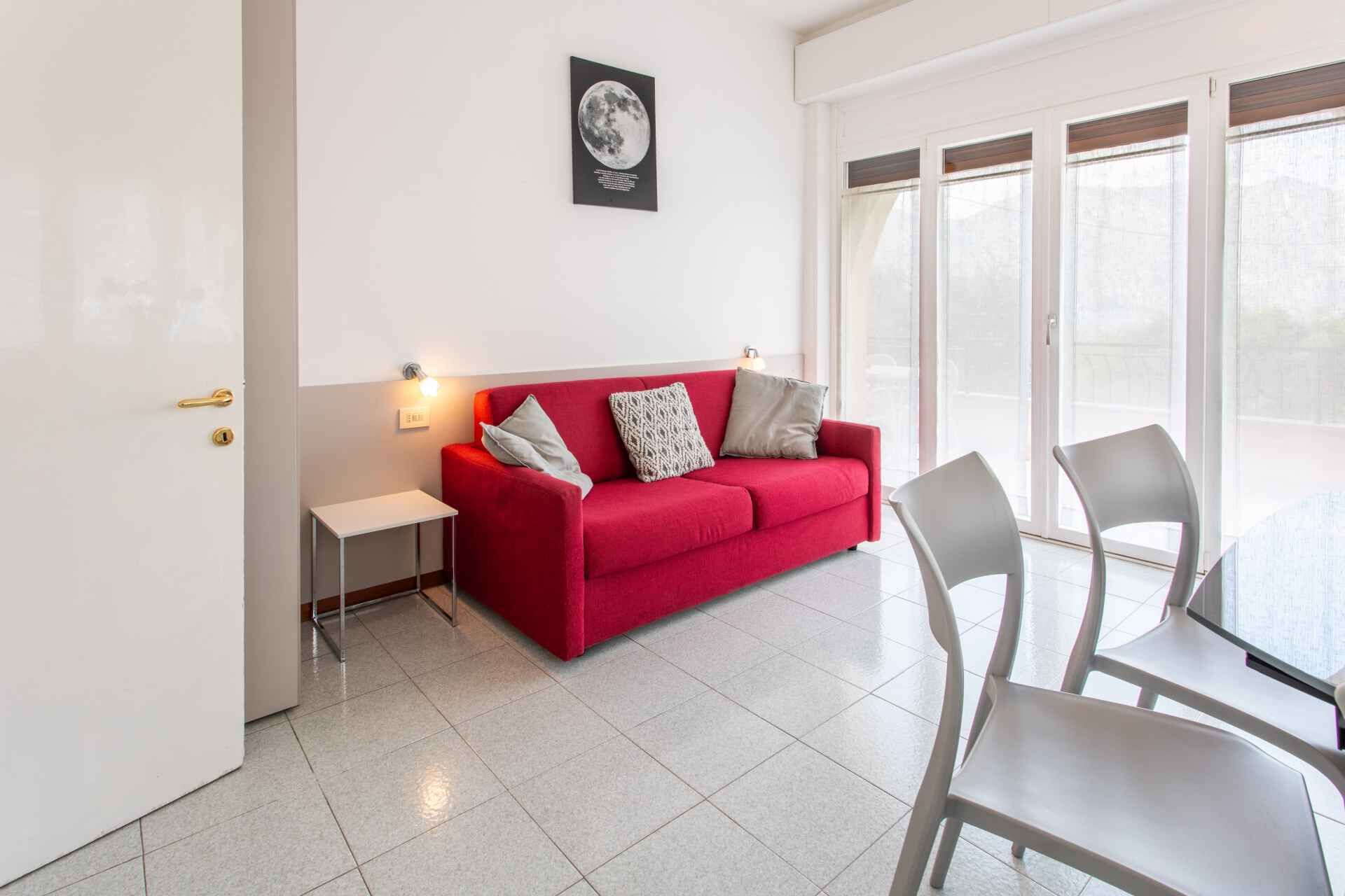 Een moderne woonkamer met een rode bank, twee grijze fauteuils, een klein wit tafeltje en een groot raam met zonwering.