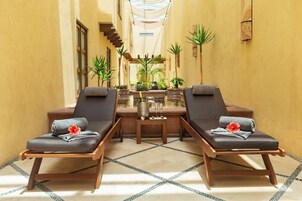 A sunlit indoor space with two lounge chairs, a small table with a drink and flowers, and a central water feature.