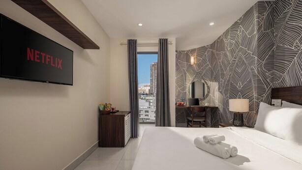 A hotel room with a large bed, a TV mounted on the wall, a desk with a chair, and a view of the city through a window. - KLY Hotel Vung Tau (Ho Chi Minh City)