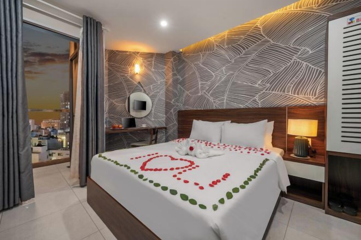 A hotel room with a large bed, a city view, a desk with a lamp, and a modern wall design.