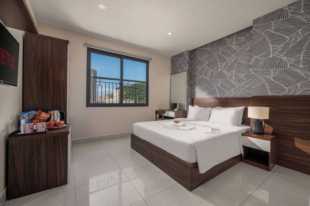 A modern hotel room with a large bed, a wooden headboard, a nightstand, a desk, and a window with a city view.