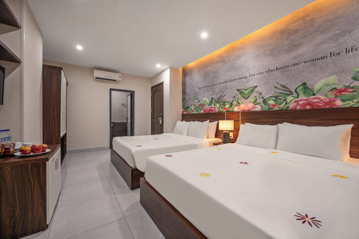 A hotel room with two beds, a wooden desk with fruit, a wall mural, and a door.