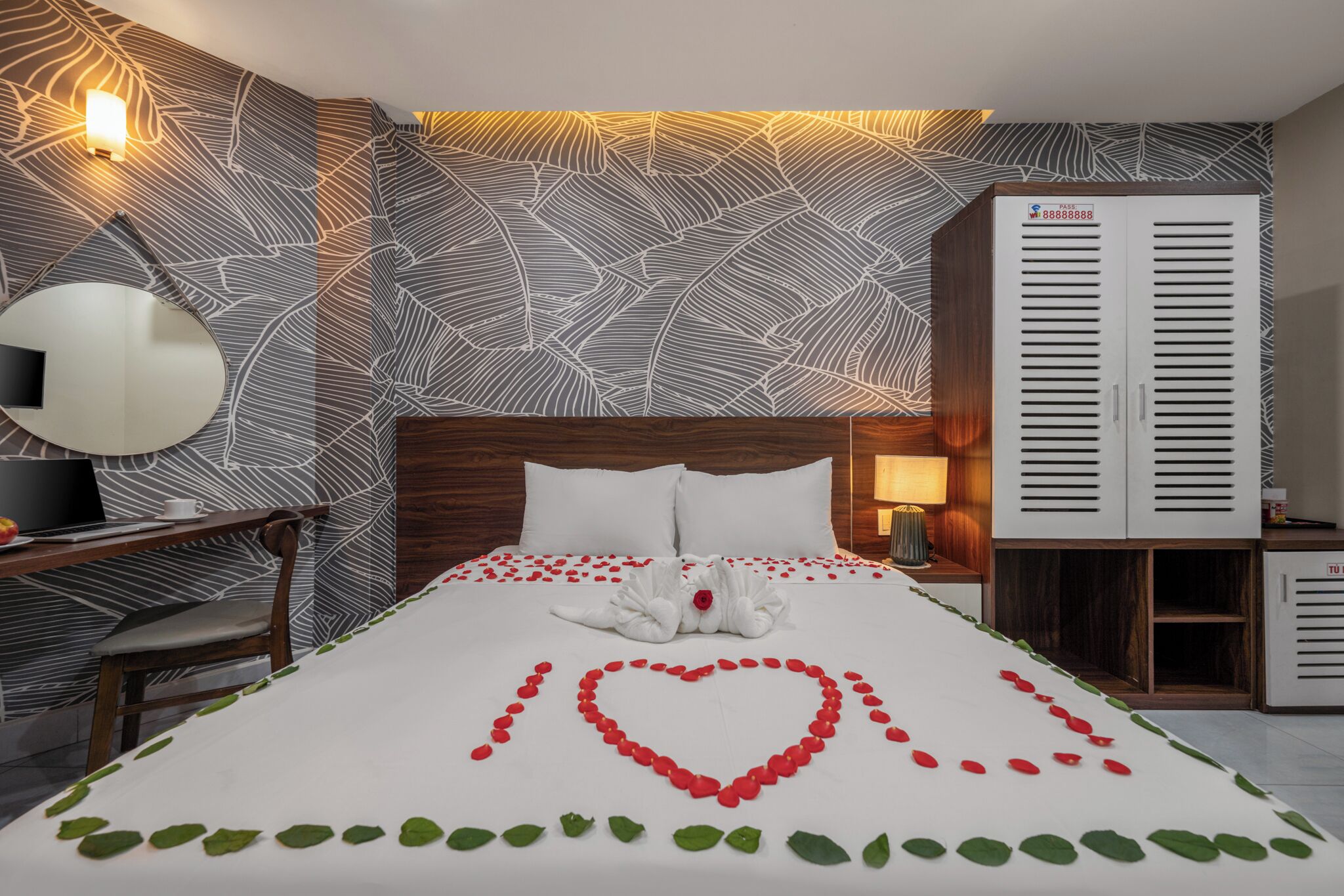 A hotel room with a large bed, a wooden headboard, a desk with a chair, a mirror, a wardrobe, and a wall with a leaf pattern.