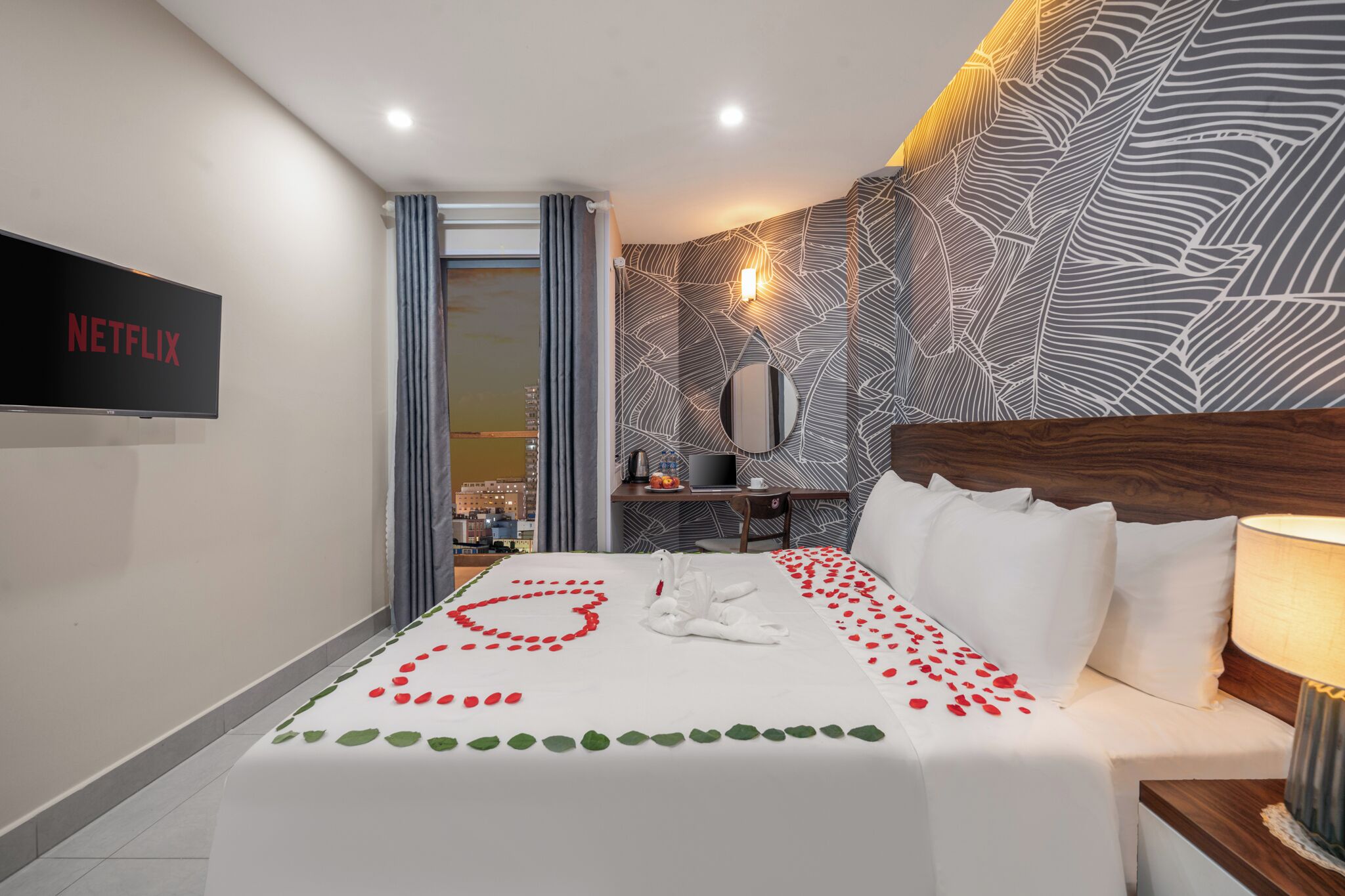 A hotel room with a bed adorned with rose petals, a flat-screen TV, a desk with a laptop, and a city view.
