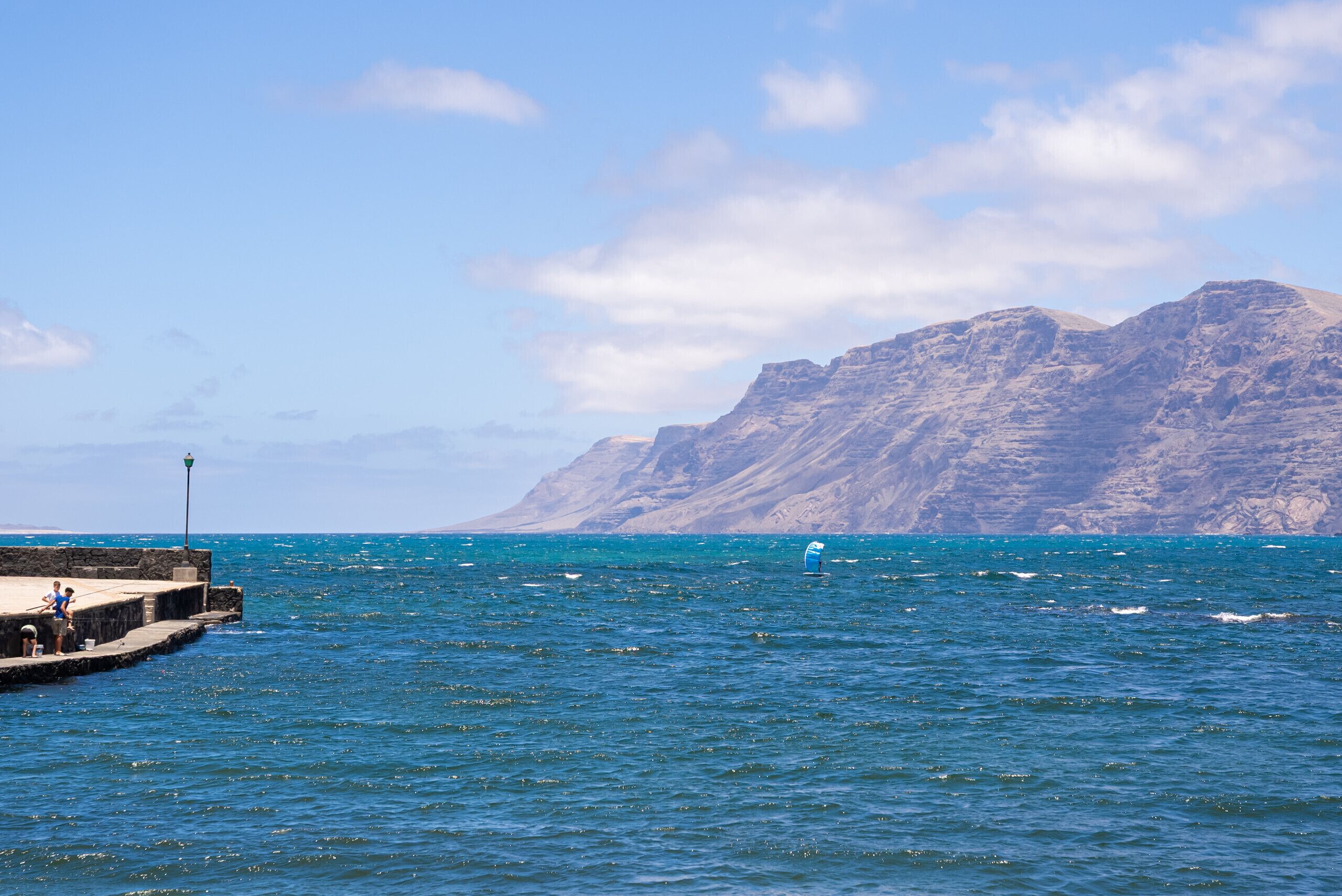 A coastal scene with a pier extending into the sea, a rocky mountain in the background, and a clear sky.