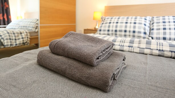A neatly made bed with a checkered pillowcase, a folded gray towel, and a wooden headboard.
