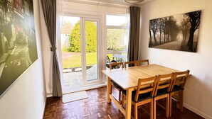 A dining area with a wooden table and chairs, a large window with curtains, and a painting on the wall.