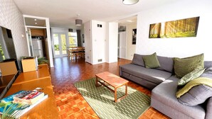 A spacious living area with a sofa, coffee table, and a dining table. There is a kitchen in the background, and the room has a modern design with a large window.