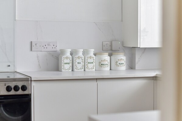 A kitchen with white cabinets, a marble backsplash, and five labeled jars on the counter. - Amazing Beachside 2Bd Family Home With Garden (Margate)