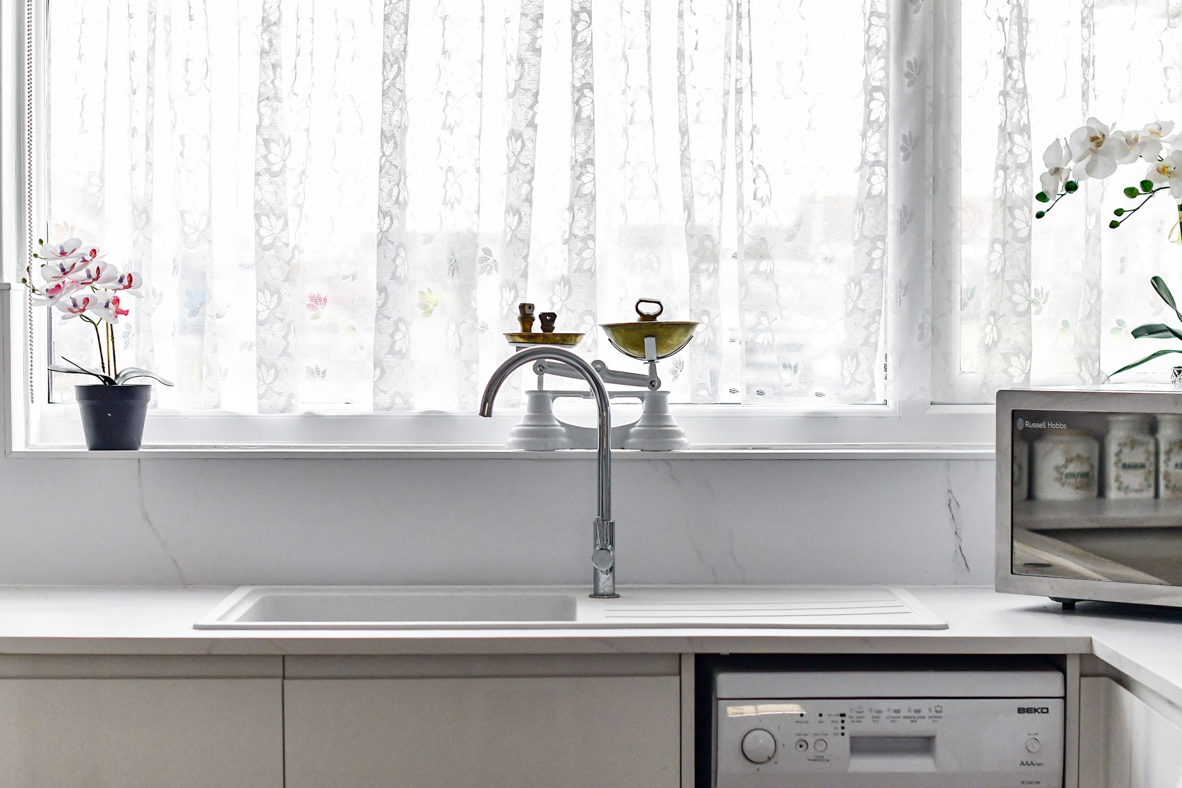A kitchen with a sink, a window with sheer curtains, a potted orchid, and a microwave.