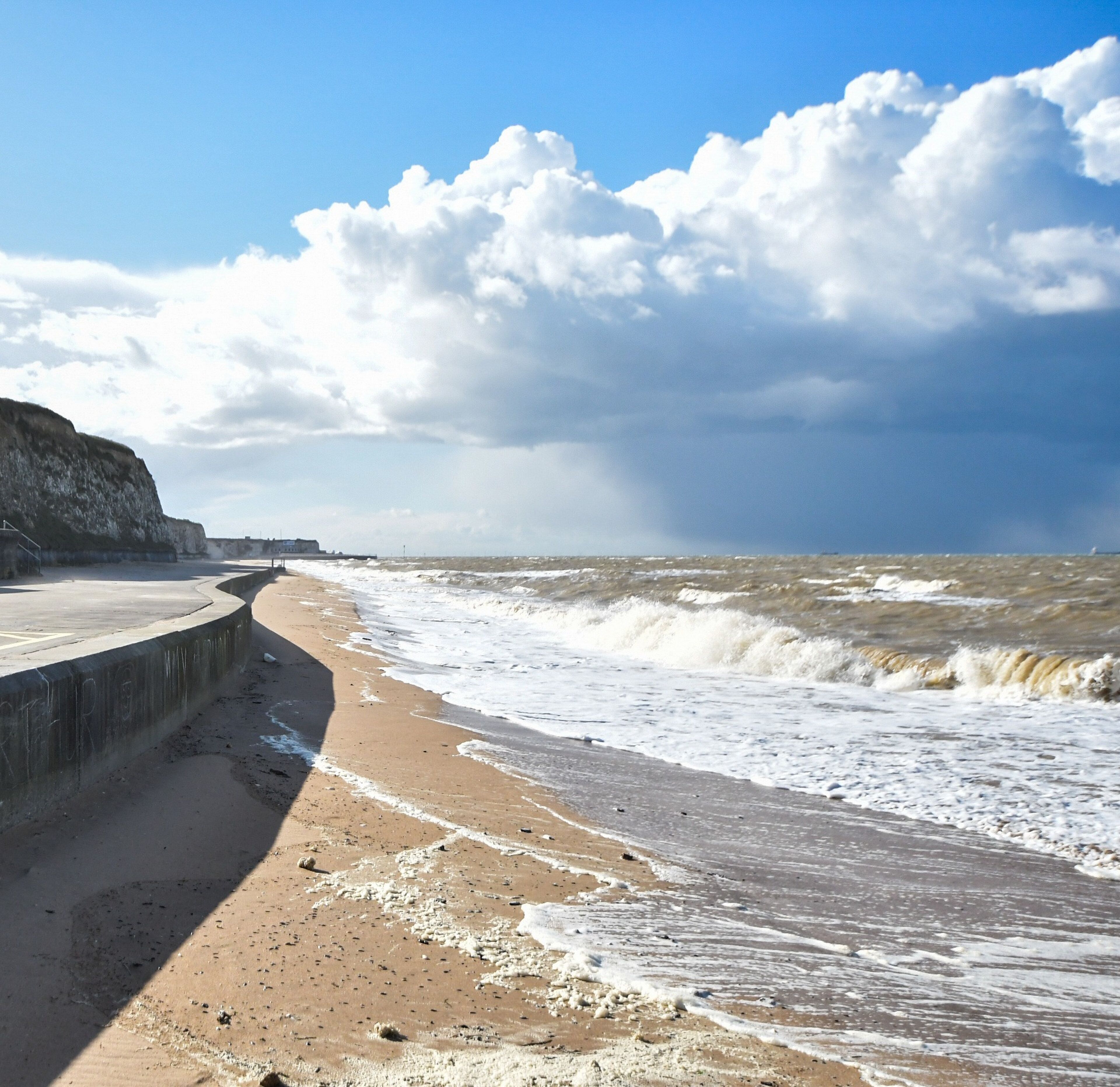 A beach with a curved concrete walkway, waves crashing onto the shore, and a cliff in the background.