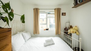 Un lit bien fait, avec du linge de lit blanc et une serviette enroulée, une fenêtre avec des rideaux beiges, une plante et des peluches sur une étagère.