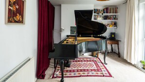 Un piano à queue noir se trouve dans une pièce où l’on aperçoit un rideau rouge, un tapis à motifs et une bibliothèque remplie de livres et d’objets décoratifs.