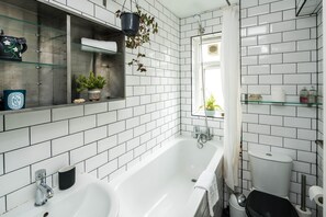 Un baño con paredes de azulejos blancos, una bañera blanca, un lavamanos y un inodoro. Hay estantes con artículos de aseo y una planta colgante.