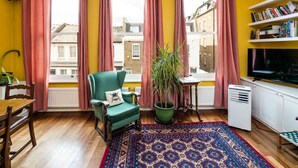 A room with a green armchair, a potted plant, a patterned rug, a radiator, a bookshelf, and a television.