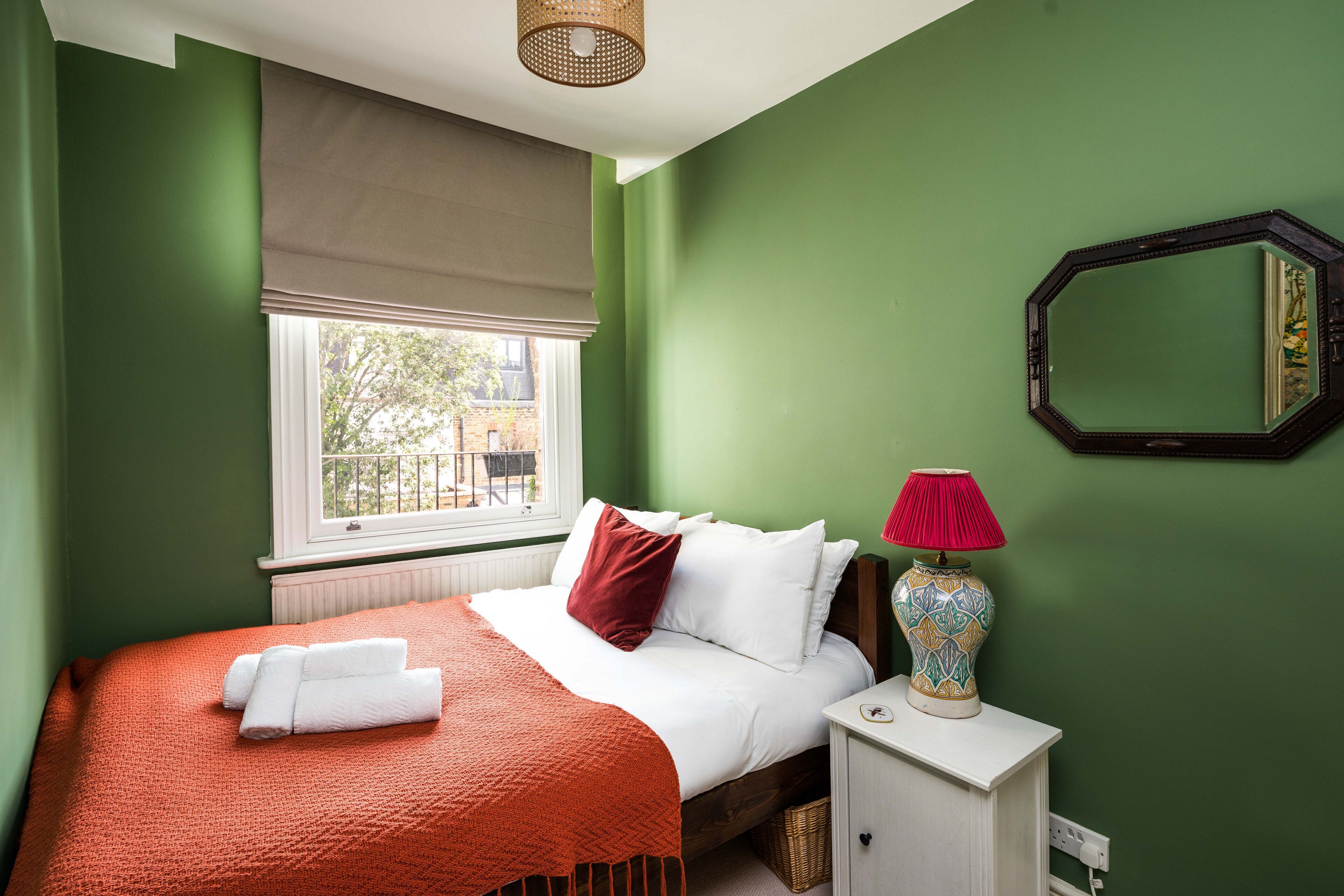 A bedroom with green walls, a bed with white bedding and an orange blanket, a bedside table with a lamp, and a window with a brown blind.