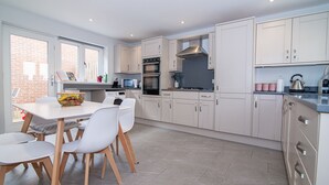 A modern kitchen with white cabinets, a dining area with a table and chairs, and a brick wall visible through the window. - Elegant Ludlow Haven: 4Bed Ultimate Tranquility (Ludlow)