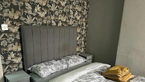 Un dormitorio con cabecera gris, dos mesitas de noche, una cama con ropa de cama floral y un papel tapiz estampado.