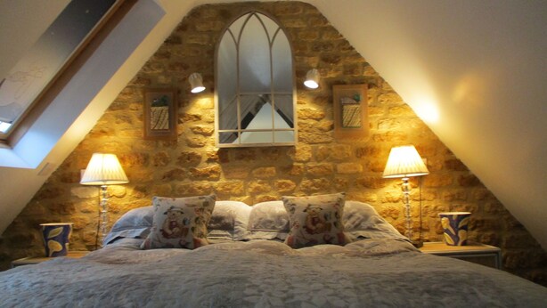 A bedroom with a stone wall, a skylight, a bed with floral bedding, and two bedside lamps. - Wonderful Picturesque Countryside Cottage (Banbury)