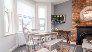A dining area with a table and chairs, a fireplace, and a television on the wall. - Little Boat: A Modern Beachside Flat (Bournemouth)
