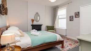 A neatly arranged bedroom with a bed, bedside lamp, a small table, a chair, a fireplace, and a window with curtains. - Little Boat: A Modern Beachside Flat (Bournemouth)