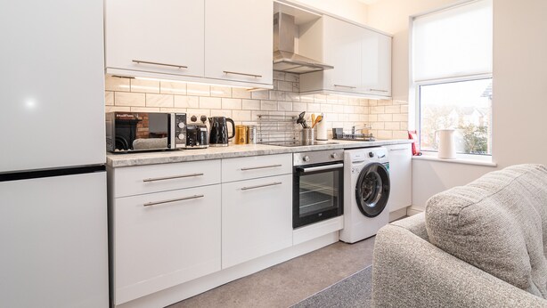 A modern kitchen with white cabinetry, a built-in oven, a washing machine, and a window providing natural light. - Little Boat: A Modern Beachside Flat (Bournemouth)