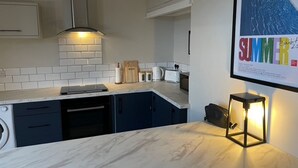 A modern kitchen with dark blue cabinets, a white countertop, and a built-in dishwasher. There is a framed poster on the wall and a small table with a lamp.