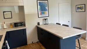 A modern kitchen with a central island, dark cabinetry, and a white countertop. There are framed artworks on the walls and a door to the right.