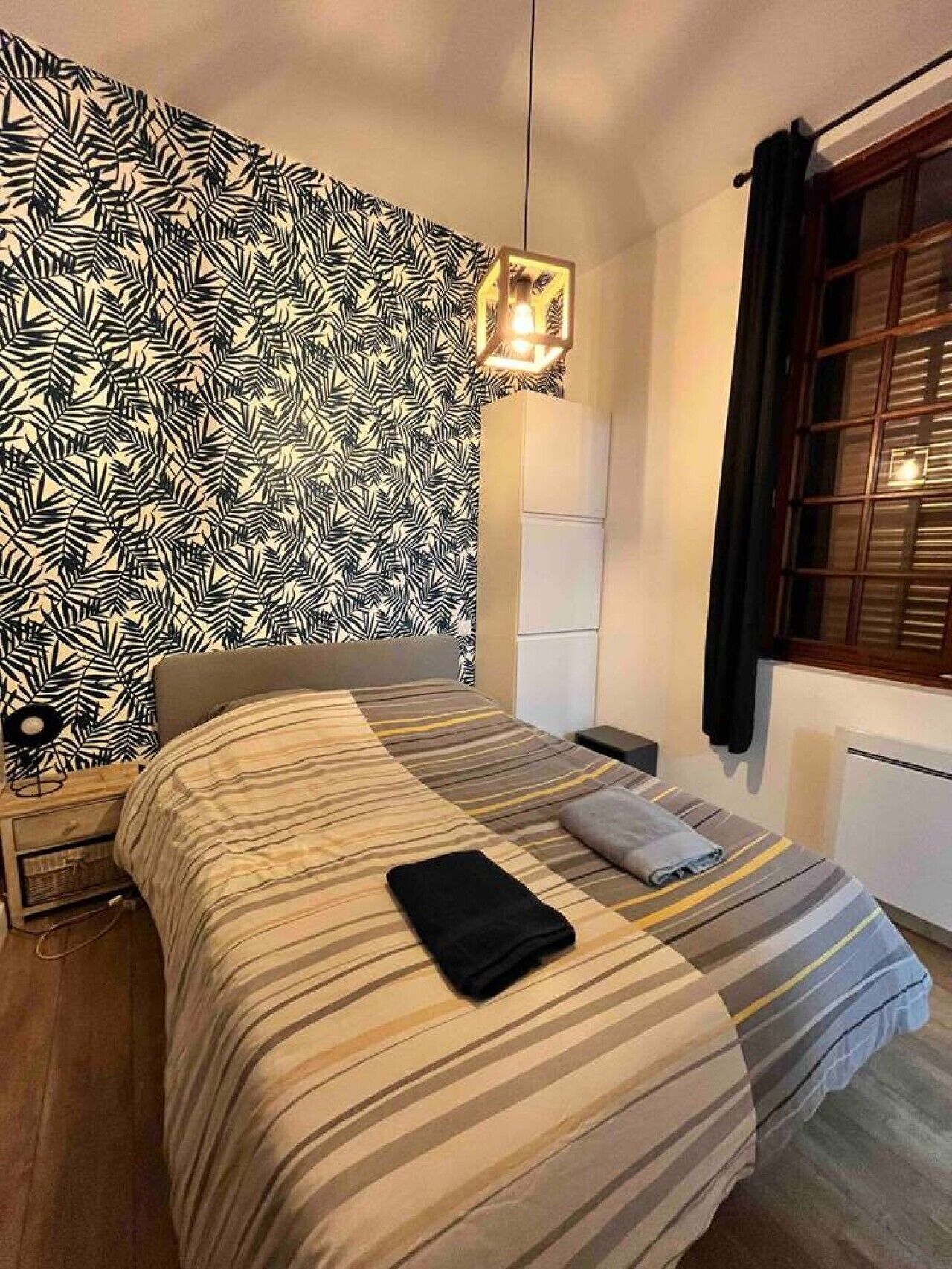 Une chambre à coucher avec un lit, une table de chevet, une lampe fixée au mur et une fenêtre avec des rideaux.