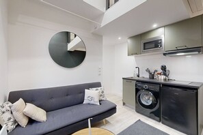 Un espacio habitable compacto con un sofá gris, lavadora, microondas y una pequeña cocina.