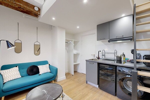 Un espacio habitable compacto con kitchenette, un sofá, lavadora y techo con vigas a la vista.