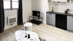 A modern kitchen with a built-in microwave, a sink, and a dishwasher. There is a round marble table with candles and a small black side table. The room has a large window with curtains and a radiator.