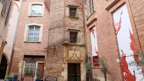 A narrow alleyway with a tall brick building, potted plants, and a bench.
