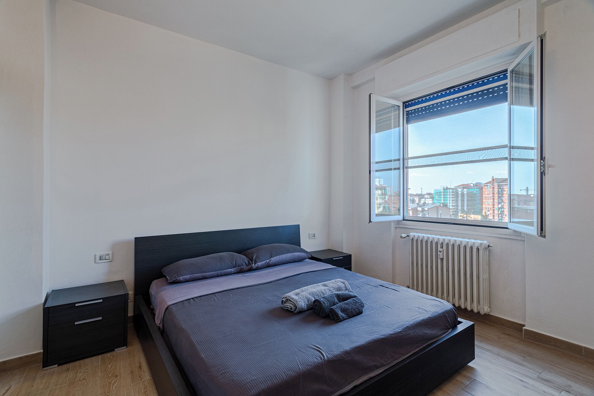 Ein Schlafzimmer mit Bett, Nachttisch, einem Fenster mit Jalousien und einer Heizung.